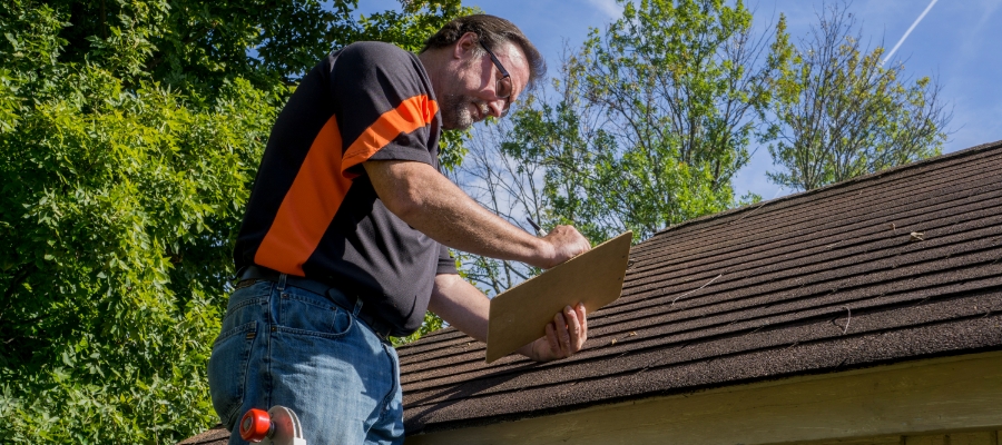 man on a ladder writes on clipboard near a roof